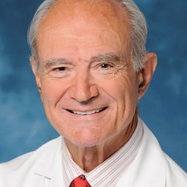 Dr. George Andros smiles in a white lab coat and red tie against a sky blue background