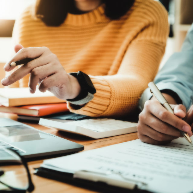 Two people collaborating over contract, books, calculatory, and laptop