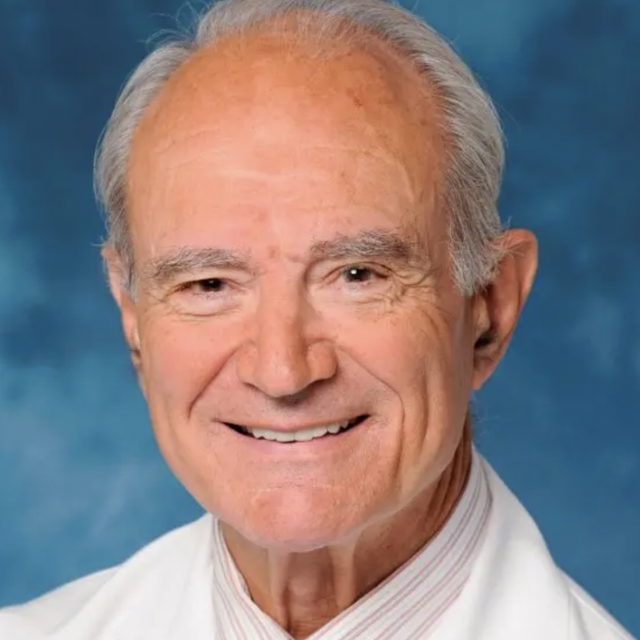 Dr. George Andros smiles in a white lab coat and red tie against a sky blue background